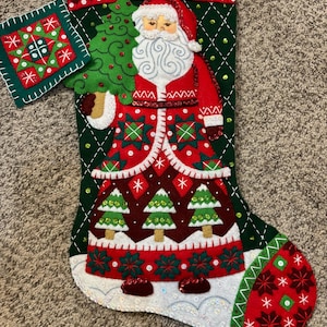 May include: A Christmas stocking featuring Santa Claus holding a green Christmas tree. The stocking is red, green, and white with decorative patterns and sequins. A matching square ornament is attached.