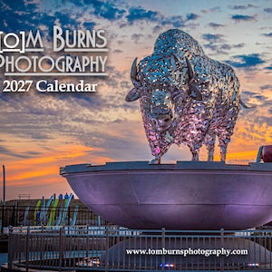 May include: A 2027 calendar featuring a metallic, textured bison sculpture standing atop a large, circular basin against a sunset sky. The text reads Tom Burns Photography 2027 Calendar and www.tomburnsphotography.com. The scene includes a metal fence.