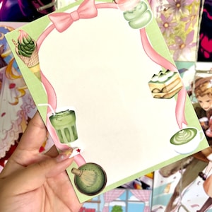 May include: A rectangular sheet of light green stationery paper featuring a pink ribbon border. The design includes illustrations of matcha-themed treats, including a soft-serve cone, a layered cake, a glass of iced tea, a latte, and a bamboo whisk.