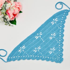 May include: A light blue crocheted shawl with dragonfly cutouts. The triangular shawl has a scalloped edge and a looped tie. The handmade accessory is displayed on a white surface, with pink and white flowers in the background.