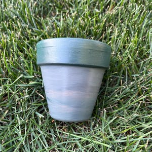 May include: A small, green and gray terracotta pot with a white rim. The pot is sitting on a bed of green grass.