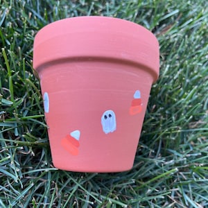 May include: A small terracotta flower pot painted with white ghosts and orange and white candy corn designs.