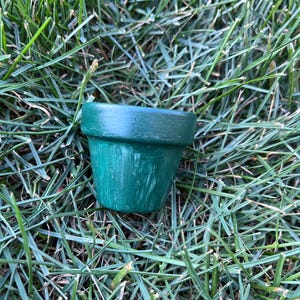 May include: A small, green terracotta pot sits on a bed of green grass. The pot is made of clay and has a rough, textured surface.