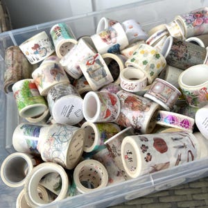May include: A selection of decorative washi tape rolls in a clear plastic container. The tape features diverse designs, including floral patterns, illustrations, and text. The colours include white, pink, green, and brown.
