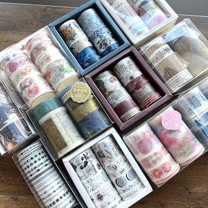 May include: Assortment of decorative washi tapes in clear plastic boxes. The tapes feature various designs, including floral patterns, vintage maps, and geometric shapes. The color palette includes blues, pinks, greens, and neutrals.
