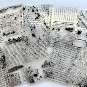 May include: A collection of clear silicone stamps featuring various designs including cursive handwriting, printed text, butterfly illustrations, ink splatters, floral motifs, a feather, and decorative borders. The stamps are transparent and flexible.