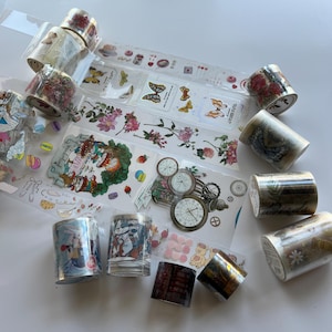 May include: A collection of decorative washi tape rolls featuring various printed designs including butterflies, floral patterns, clock faces, macarons, and whimsical character illustrations. The rolls are arranged with strips of tape unrolled to display the patterns.