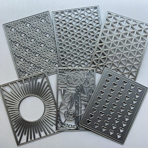 May include: A set of six rectangular metal craft dies featuring various intricate cut-out patterns including geometric grids, a sunburst design with a central circle, a floral arrangement, and a repeating grid of small heart shapes.