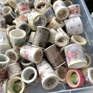May include: A selection of washi tape rolls with diverse patterns and colours. Designs include florals, landscapes, and other patterns. The rolls are stored in a clear plastic container.