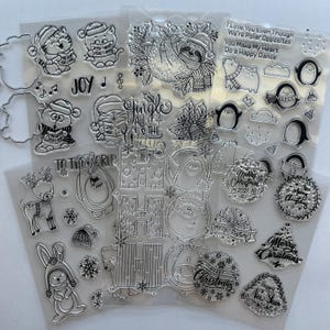 May include: A collection of clear photopolymer stamps featuring Christmas-themed illustrations including cats, a sloth, penguins, a reindeer, a rabbit, and a bear. Text includes "Jingle All The Way," "Joy," "To The World," and "Merry Christmas."