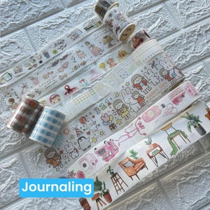 May include: A selection of decorative washi tapes featuring diverse designs such as floral motifs, cartoon characters, and geometric patterns. The tapes are presented in rolls and strips, ideal for journaling and craft activities. The word "Journaling" is in a blue box.