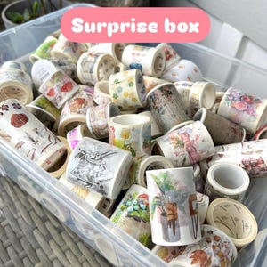 May include: A clear plastic container overflowing with decorative washi tape rolls. The tape rolls display diverse designs, such as floral patterns, illustrations, and text. The text "Surprise box" is shown in a pink banner.