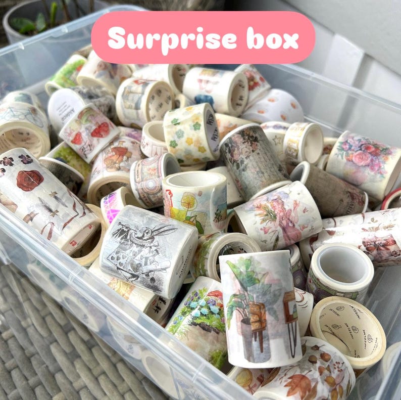 May include: A clear plastic container overflowing with decorative washi tape rolls. The tape rolls display diverse designs, such as floral patterns, illustrations, and text. The text "Surprise box" is shown in a pink banner.