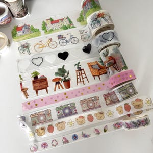 May include: A selection of decorative washi tapes with diverse designs. The tapes showcase illustrations of bicycles, hearts, cameras, bears, flowers, and houses. The colour scheme includes pink, green, yellow, and white.