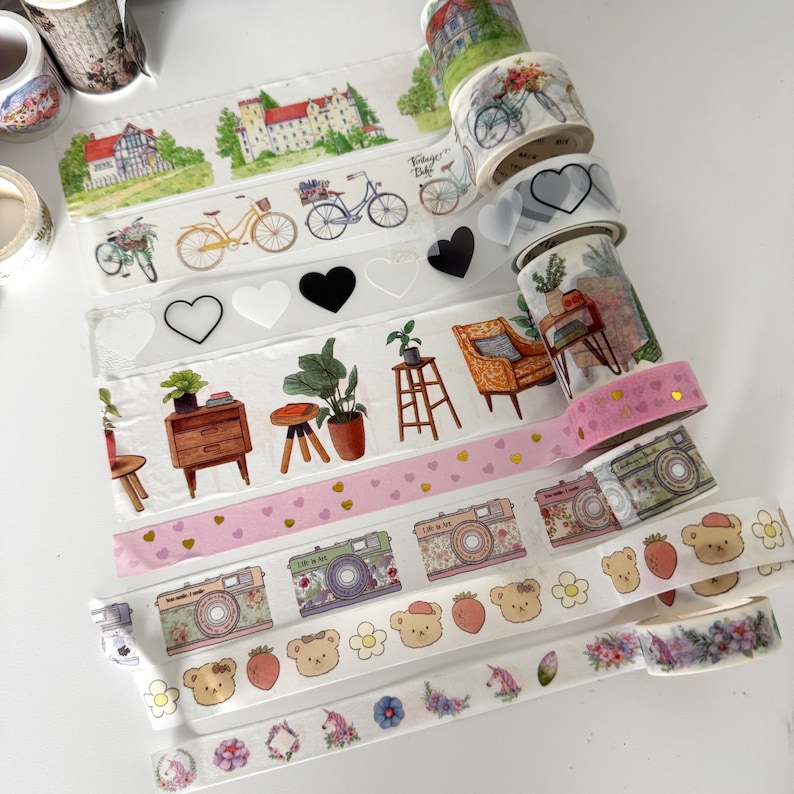 May include: A selection of decorative washi tapes with diverse designs. The tapes showcase illustrations of bicycles, hearts, cameras, bears, flowers, and houses. The colour scheme includes pink, green, yellow, and white.