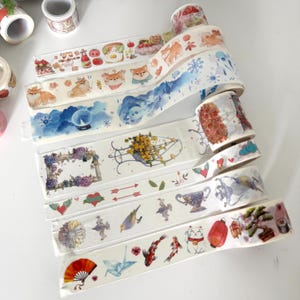 May include: A selection of decorative washi tape rolls with diverse designs. The tapes showcase illustrations of Shiba Inu dogs, floral arrangements, koi carp, and Japanese-themed imagery. The colour palette includes white, blue, red, and yellow.