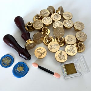 May include: A collection of brass wax seal stamps with wooden handles, featuring various holiday-themed designs. Includes blue wax seals, an eyeshadow applicator, and a small container of gold eyeshadow. The seals have Christmas and winter motifs.