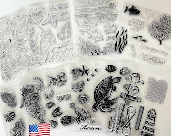 May include: Clear stamps featuring various sea-themed designs, including fish, seashells, dolphins, and a turtle. Text includes "Life is better at the Beach", "Go with the flow", "SEAFOOD", "Life Awesome", "Vacances", "Thank You", and "I'm here for you always". An American flag is also present.