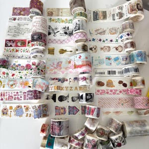 May include: A selection of decorative washi tapes in diverse designs and colours. The tapes showcase floral patterns, travel themes, and other illustrations. Some tapes have text, and the rolls are stacked on a white surface.