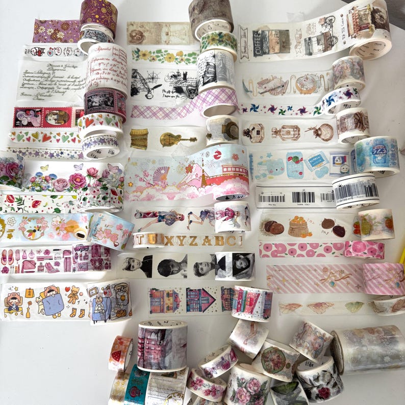 May include: A selection of decorative washi tapes in diverse designs and colours. The tapes showcase floral patterns, travel themes, and other illustrations. Some tapes have text, and the rolls are stacked on a white surface.