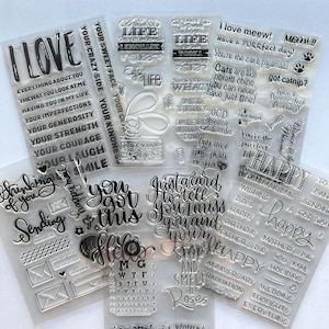 May include: A collection of clear photopolymer stamp sheets featuring various black printed phrases, cat-themed quotes, and decorative icons. The designs include text like I LOVE, YOU GOT THIS, HAPPY, and various holiday greetings, alongside small envelopes and paw prints.