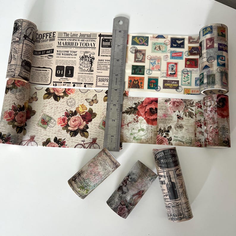 May include: Several rolls of decorative washi tape with vintage designs. The tape features floral patterns, newspaper clippings, postage stamps, and text like "Coffee Shop" and "Married Today." A metal ruler is placed next to the tape.