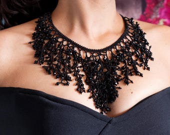 May include: A black beaded necklace with a cascading design. The necklace features intricate beadwork and a delicate, fringed appearance. The jewelry is worn against a black top, creating a striking contrast.