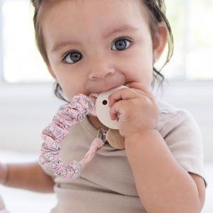 May include: A baby wearing a light brown onesie is holding a pink floral pacifier and a beige wooden teether in their mouth. A small, beige knitted doll with a pink dress is in the baby's lap.