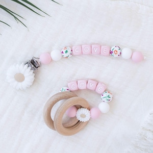 May include: A baby accessory set featuring a pacifier clip and a wooden teether. The pacifier clip and teether have pink, white, and floral beads spelling "COVE". Both items include a white daisy flower and are made of wood and silicone.