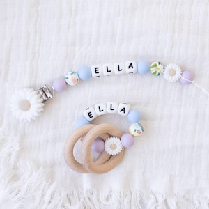 May include: A baby pacifier clip and teether set. The clip and teether feature the name "ELLA" spelled out with white letter beads. The clip has light blue and floral beads, a daisy flower, and a metal clip. The teether has wooden rings and matching beads.