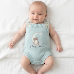 May include: A light blue baby romper with a teddy bear and the name "Christopher" embroidered on the front. The romper has wooden buttons on the straps and is made of a soft, knitted material. The baby is lying on a white sheet.