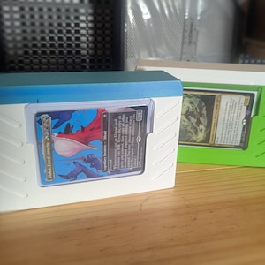 May include: Two card storage boxes. One is white with a blue top, the other is white with a green top. Each box holds a trading card. The cards have colorful illustrations and text. The boxes are on a wooden surface.