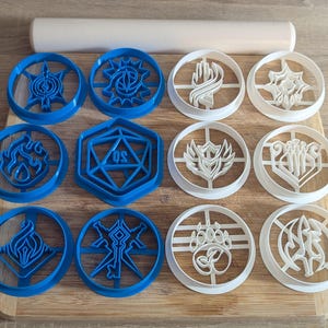 May include: A collection of blue and white cookie cutters in various geometric and symbol designs, including a 20-sided die. A rolling pin rests above the cutters on a wooden cutting board. The cutters are likely for baking.