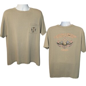 以下が含まれることがあります： 左側にポケットが付いた、ベージュ色の半袖Tシャツ。ポケットには十字のデザインがあります。Tシャツの背面には、"Indianapolis Harley-Davidson Indiana"と書かれたワシのデザインがあります。