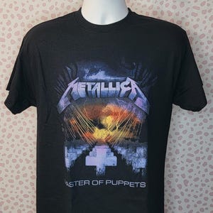 May include: Black t-shirt featuring the band name "Metallica" in a silver and purple font above a graphic design. The graphic includes a cross-shaped structure and the words "Master of Puppets" in white.