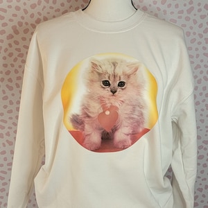 May include: White sweatshirt featuring a graphic of a fluffy kitten holding a pink heart. The kitten has green eyes and is surrounded by a yellow circle.