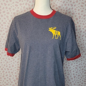 May include: A grey t-shirt with a red collar and sleeve trim. A yellow moose graphic with the number 92 is on the front. The shirt is displayed on a mannequin.