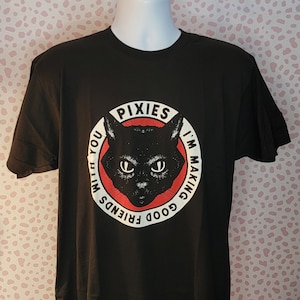 Könnte beinhalten: Schwarzes T-Shirt mit einem kreisförmigen Grafikdesign, das eine schwarze Katzenillustration zeigt. Die Grafik enthält den Bandnamen "PIXIES" und den Satz "I'M MAKING GOOD FRIENDS WITH YOU". Das Design ist in Weiß und Rot gehalten.