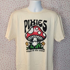 Puede incluir: Una camiseta de color crema con un gráfico en blanco y negro de un hongo con manchas rojas y el texto "Pixies" encima. El texto "Where is my mind?" está debajo del hongo.