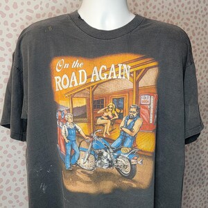 Peut inclure: T-shirt gris fonc&eacute; avec un motif repr&eacute;sentant une moto, une station-service et le texte "On the Road Again". Le motif comprend des personnages et un distributeur de soda vintage. Le t-shirt a un aspect us&eacute;.