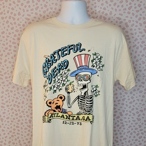 May include: A cream-colored t-shirt with a graphic of a skeleton wearing a top hat and sunglasses, holding an apple and standing next to a bear. The text "Grateful Dead" is written in blue and the text "Atlanta, GA 12-12-73" is written in black.