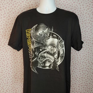 Puede incluir: Camiseta negra con un estampado gráfico de una banda de metal, Iron Maiden, con una calavera y un ojo de cyborg. El nombre de la banda está escrito verticalmente en amarillo en el lado izquierdo de la imagen.