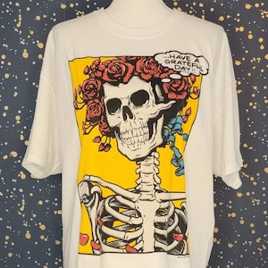 May include: White t-shirt with a graphic print of a skeleton with a crown of red roses. The skeleton is smiling and has a speech bubble that says "...have a grateful day!"