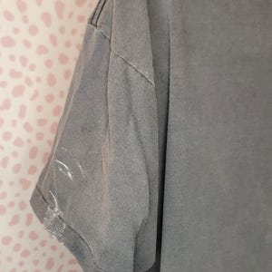 Peut inclure: Gros plan d'un t-shirt gris &agrave; manches courtes. Le t-shirt a un aspect d&eacute;lav&eacute; et des coutures visibles le long des coutures. Il y a des marques de peinture blanche sur la manche. Le fond est un mur rose &agrave; pois blancs.