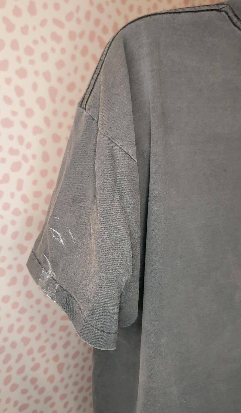 Peut inclure: Gros plan d'un t-shirt gris &agrave; manches courtes. Le t-shirt a un aspect d&eacute;lav&eacute; et des coutures visibles le long des coutures. Il y a des marques de peinture blanche sur la manche. Le fond est un mur rose &agrave; pois blancs.