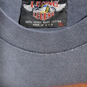 Peut inclure: Gros plan d'un t-shirt gris XL avec une &eacute;tiquette noire. L'&eacute;tiquette indique "LIVING LEGEND" avec un motif de moto et "100% HEAVY BIKER COTTON MADE IN U.S.A."