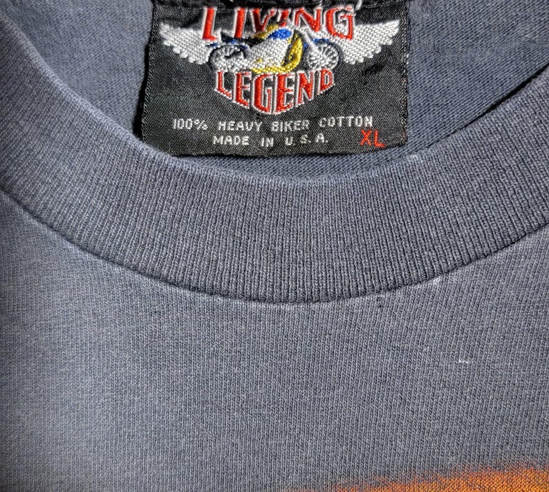 Peut inclure: Gros plan d'un t-shirt gris XL avec une &eacute;tiquette noire. L'&eacute;tiquette indique "LIVING LEGEND" avec un motif de moto et "100% HEAVY BIKER COTTON MADE IN U.S.A."