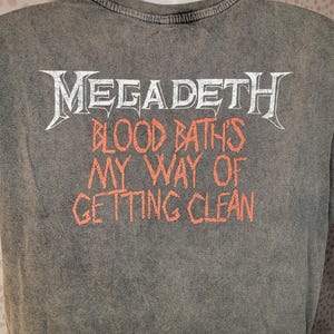 May include: Gray t-shirt with the band name "Megadeth" in white, stylized lettering. Below, in orange letters, the text reads "Blood Baths My Way of Getting Clean". The shirt has a vintage, washed appearance.