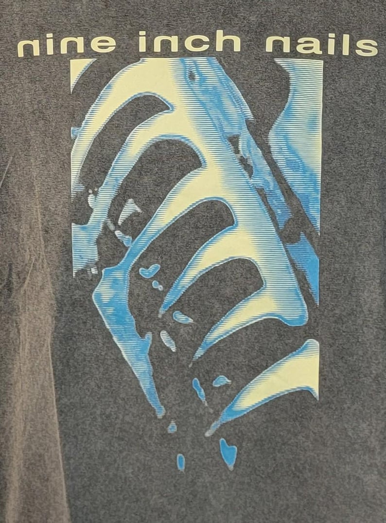 May include: A gray t-shirt featuring the band name "nine inch nails" in white block letters. The shirt has a graphic design of a blue and yellow skeletal structure. The design is centered on the shirt.