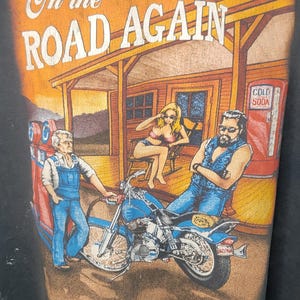 Peut inclure: T-shirt noir avec un motif repr&eacute;sentant une moto, une station-service et trois personnages. Le texte "On the Road Again" est imprim&eacute; en haut. L'image a un style vintage et r&eacute;tro.